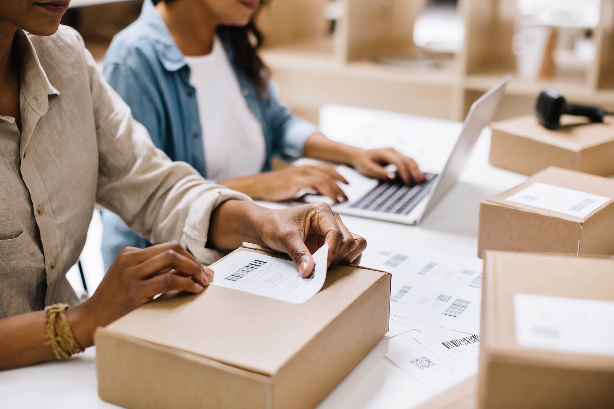 Two people preparing an e-commerce order: a shipping label applied to a parcel, laptop and boxes ready to ship
